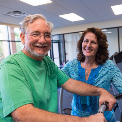 Caucasian Mature couple at the gym, he is working out white his wife is watching him smiling