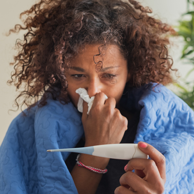 Woman wrapped in blue blanket holding a white tissue to her nose and looking at a thermometer